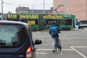 PKW, rechts daneben ein wartender Radler und eine Straßenbahn im Hintergrund.