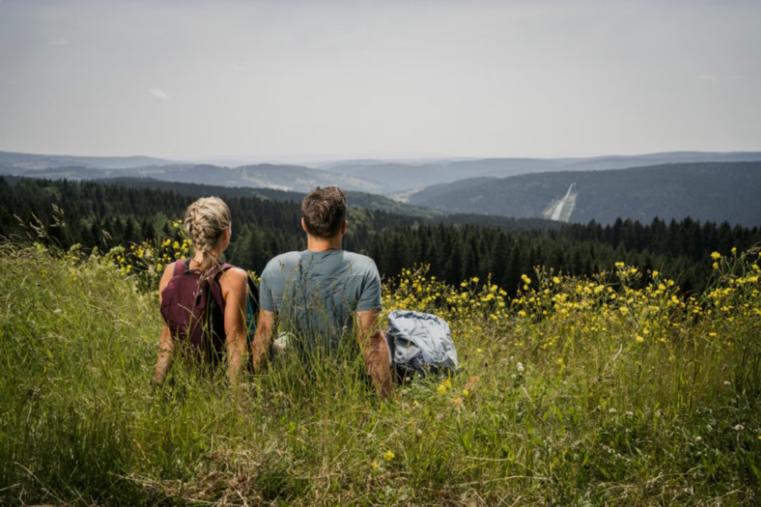 Rückenansicht von Mann und Frau, beide sitzen auf einer Bergwiese und blicken in die Ferne.