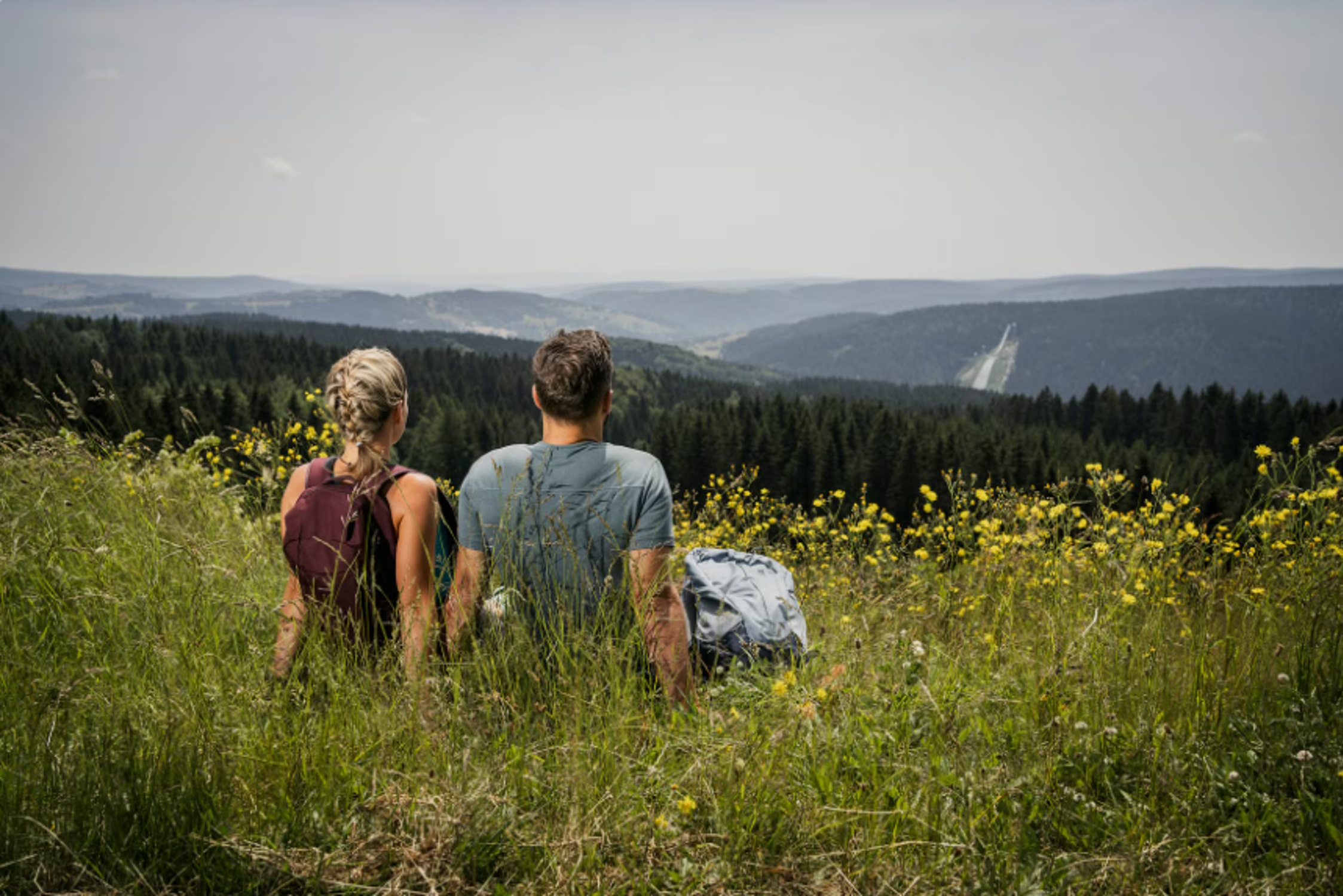 Rückenansicht von Mann und Frau, beide sitzen auf einer Bergwiese und blicken in die Ferne.