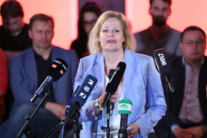 Nancy Faeser an Mikrofonen, dahinter verschwommen Personengruppe.