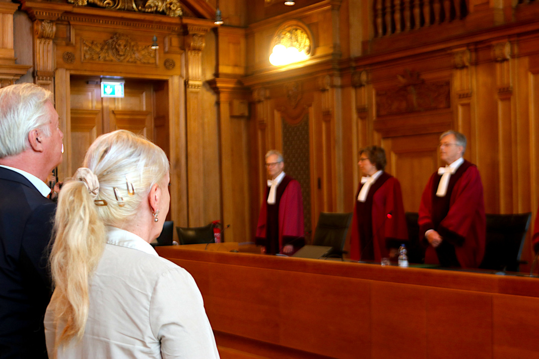 Zwei Personen, Rückenansicht im Gerichtssaal, im Hintergrund Richter in roten Roben.