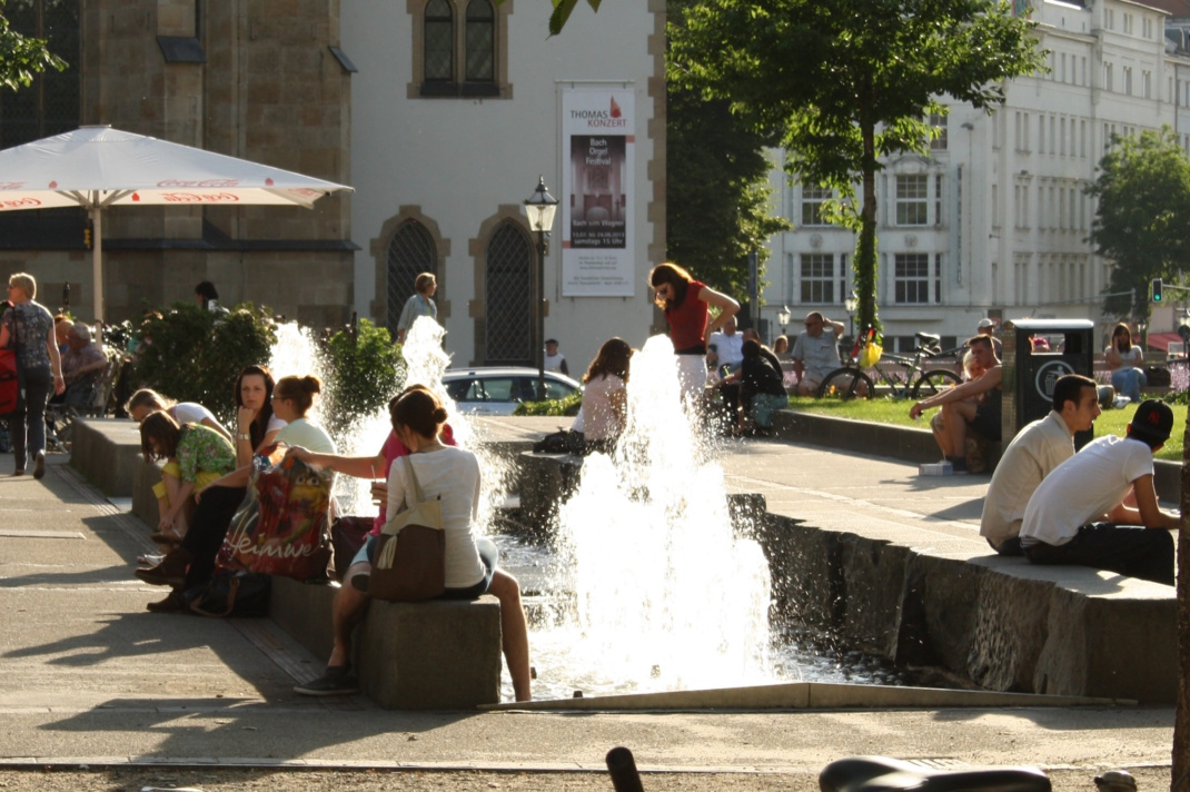 Personen in der Sonne am sprudelndem Wasser eines Brunnens.