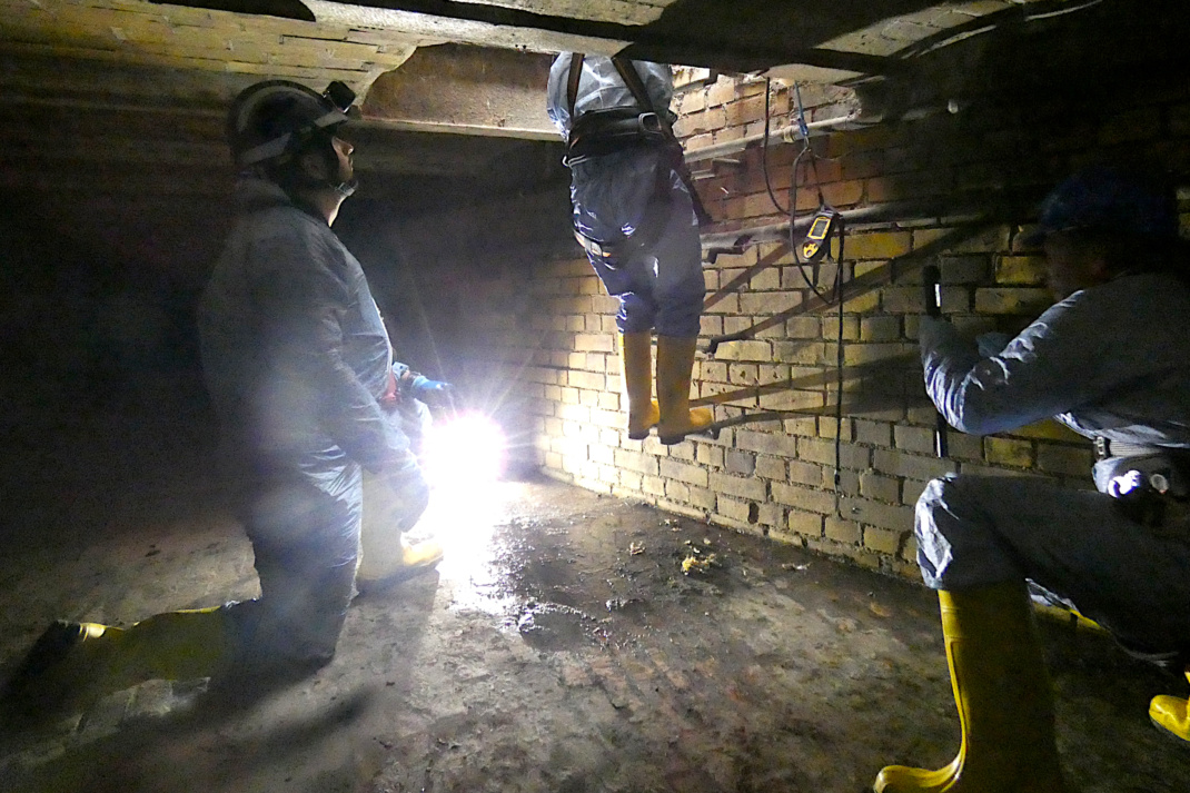 Eine Person im Schutzanzug hangelt sich in die Kanalisation hinab, daneben zwei weitere Personen.