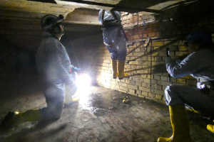 Eine Person im Schutzanzug hangelt sich in die Kanalisation hinab, daneben zwei weitere Personen.