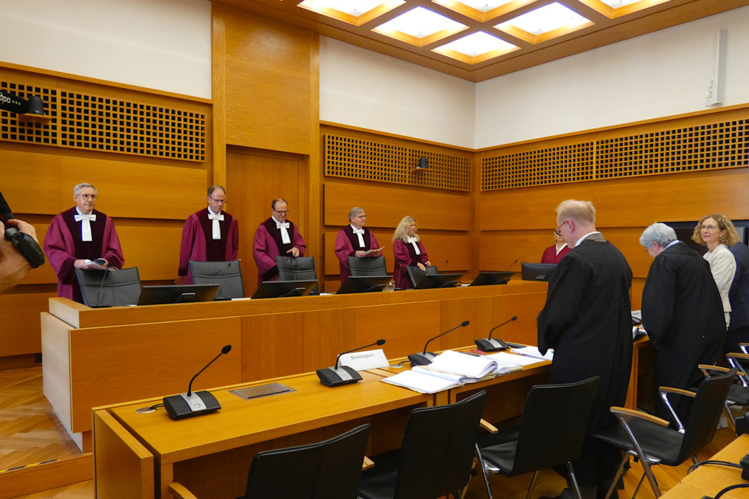 Richter in roten Roben stehen im Gerichtssaal hinter der Bank und schauen entgegen der Kamerarichtung.