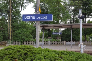 Haltepunktschild für die S-Bahn an einem Pfahl, dahinter Zaun und Überdachung.