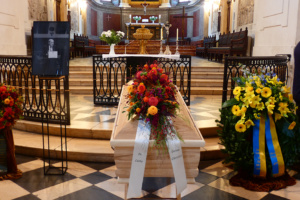 Sarg, der zur Trauerfeier in einer Kirche steht, mit Bildnis und Blumenschmuck.
