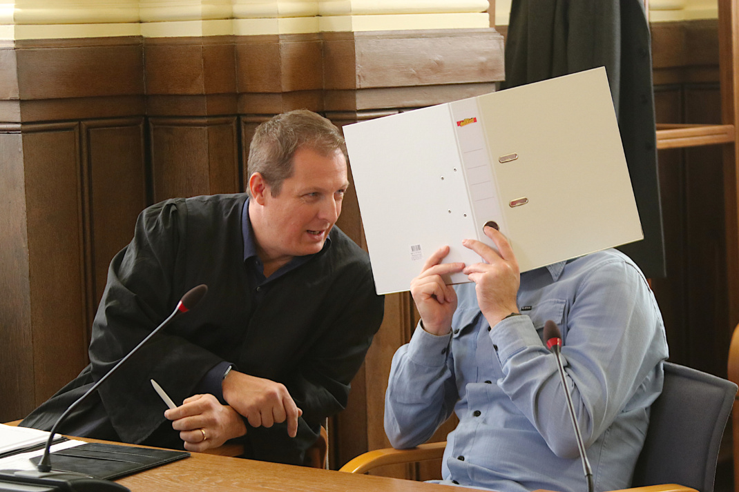 Angeklagter, der sich einen weißen Ordner vor das Gesicht hält, spricht mit seinem Anwalt im Gerichtssaal.