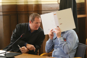 Angeklagter, der sich einen weißen Ordner vor das Gesicht hält, spricht mit seinem Anwalt im Gerichtssaal.