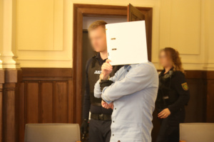 Angeklagter mit Sichtschutz betritt mit Wachtmeistern Gerichtssaal.