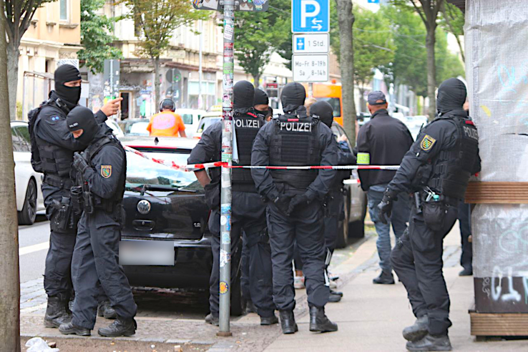 Großteils vermummte Polizisten stehen gruppiert hintere einer Absperrung.