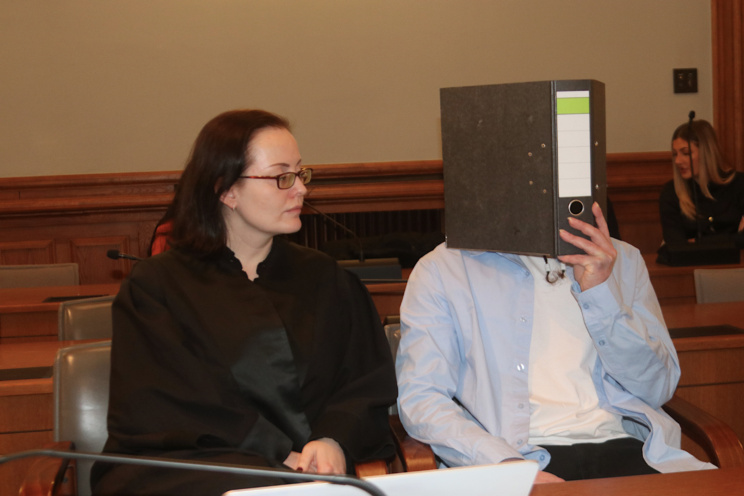 Angeklagter sitzt mit Ordner vor dem Gesicht neben seiner Verteidigerin.