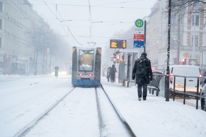 Schneefell an Haltestelle, Tram von hinten bei der Abfahrt, PKW auf separater Straße.
