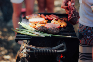 Grill in Nahaufnahme mit Grillgut, daneben andeutungsweise Menschen.