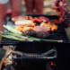 Grill in Nahaufnahme mit Grillgut, daneben andeutungsweise Menschen.