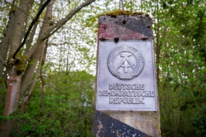 Ein Grenzpfeiler in einem Wald. Auf dem Pfeiler ist ein Schild mit der Aufschrift „Deutsche Demokratische Republik" und dem Embleme der DDR bestehend aus Hammer, Sichel und Ehrenkranz angebracht Alter DDR-Grenzpfeiler in einem Wald. Foto: Colourbox